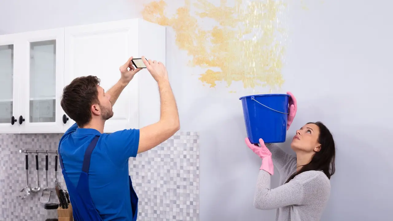 Homeowner photographing a water stained wall while another person holds a bucket under a leak