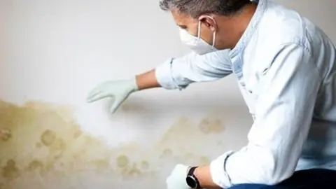 Person in a mask and gloves inspecting a water stain on a wall